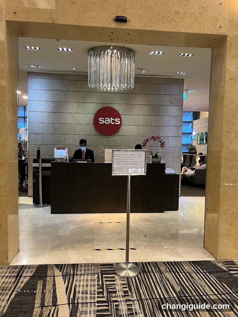 Interior view of a reception desk with staff wearing masks, behind which is the SATS logo on a tiled wall. A chandelier hangs above. A sign stands in front of the desk with text about safety measures. The floor has patterned tiles and carpeting.