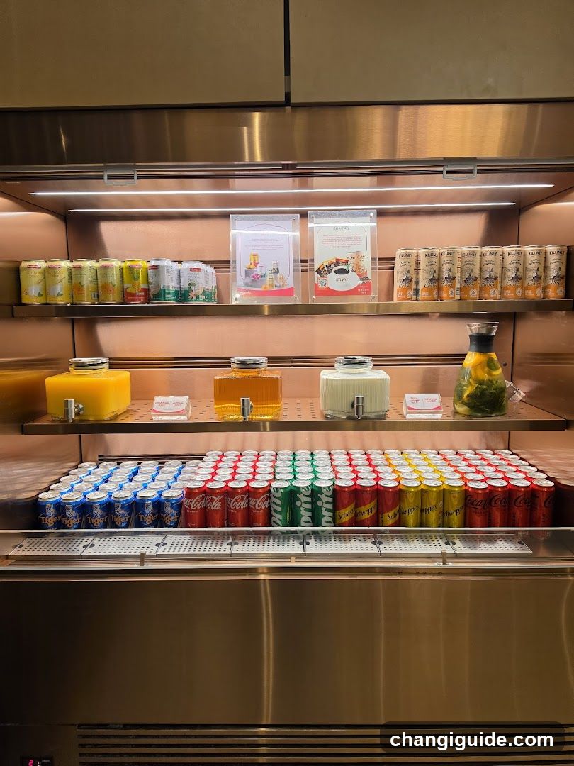 A stainless steel beverage refrigerator stocked with various canned drinks and juice dispensers. The top shelf holds cans of orange, apple, and other fruit juices alongside packaged tea or coffee products. Below are four glass dispensers filled with yellow, amber, white, and green liquids (likely juices or teas). The bottom section contains neatly arranged rows of soda cans: blue Diet Coke, red Coca-Cola, green Sprite, yellow Fanta, and another red variant.