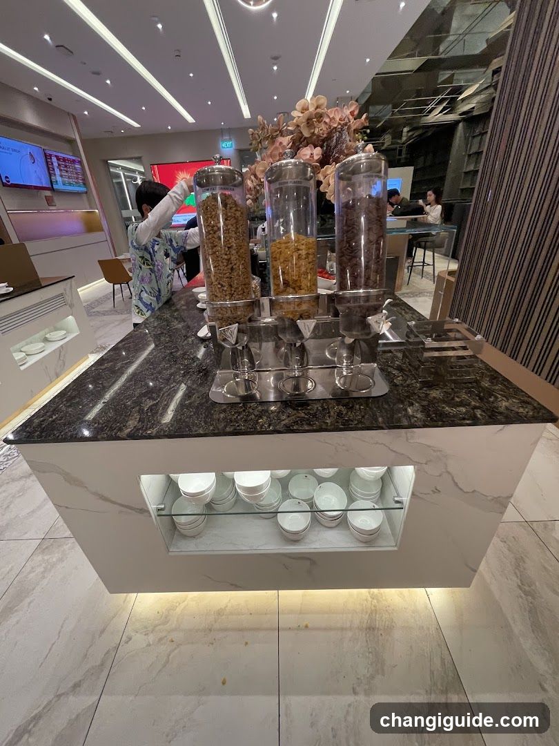 A modern breakfast buffet setup featuring three transparent cereal dispensers filled with different types of cereals on a marble countertop. Below the counter, neatly stacked white bowls and plates are illuminated by under-cabinet lighting. In the background, there is a digital menu board displaying information, along with people seated at tables enjoying their meal. The setting appears to be part of a hotel or restaurant lobby.