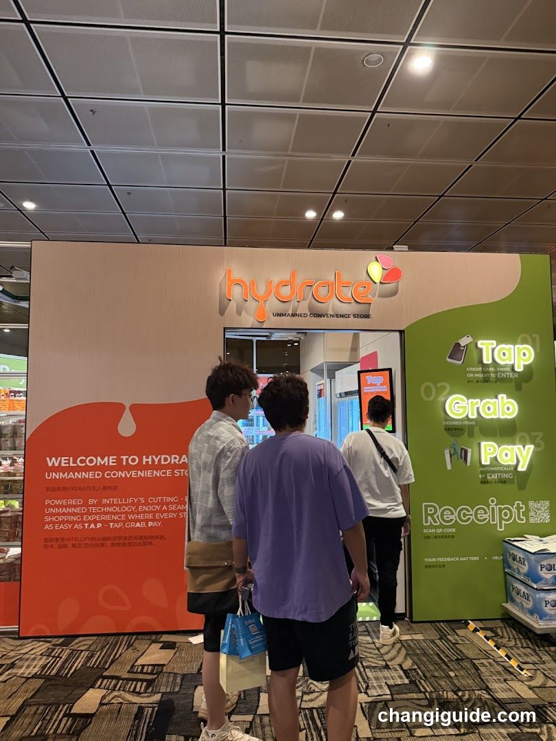 Photo of the storefront facade showing a large sign with 'hydrate' logo and colorful droplet graphics. Three people are standing in front, looking at the entrance. A green panel on the right displays instructions: Tap, Grab, Pay, Receipt. An orange section welcomes customers to Hydrate's unmanned convenience store powered by Intellify's technology.