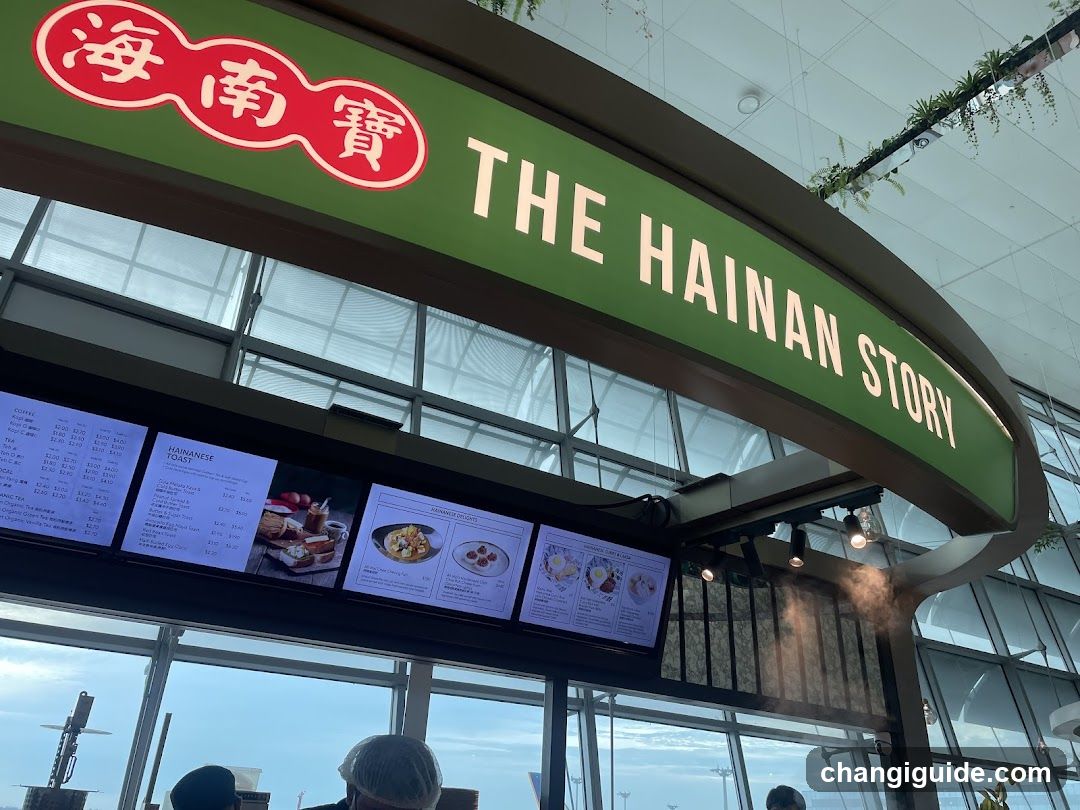 A storefront with a green sign displaying 'THE HAINAN STORY' and Chinese characters in red circles. Below the sign, there are digital screens showing menu items and prices.
