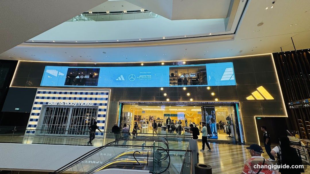 The image shows the exterior of several shop fronts in a modern shopping mall. Visible brands include Bath & Body Works, Mercedes-Benz, and Adidas. The storefronts feature illuminated signage and glass entrances with people walking by.