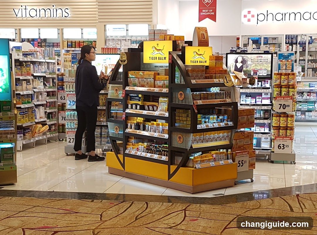 Interior view of a pharmacy or drugstore with shelves stocked with vitamins and health products. A central display features Tiger Balm products, including ointments and creams. Shelves in the background hold various packaged goods, with price tags visible. A person is browsing near the shelves. The store has bright lighting and clean, organized aisles.