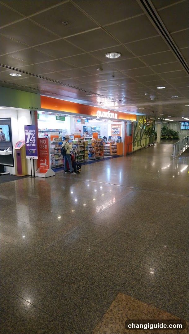 A wide shot of an airport terminal hallway featuring a brightly lit convenience store named 'Guardian' on the left. A customer stands near the entrance with luggage. To the far left, there is a digital display and promotional signage advertising a 17% GST rebate. The floor is polished granite reflecting overhead lights. In the background, greenery and directional signs are visible.