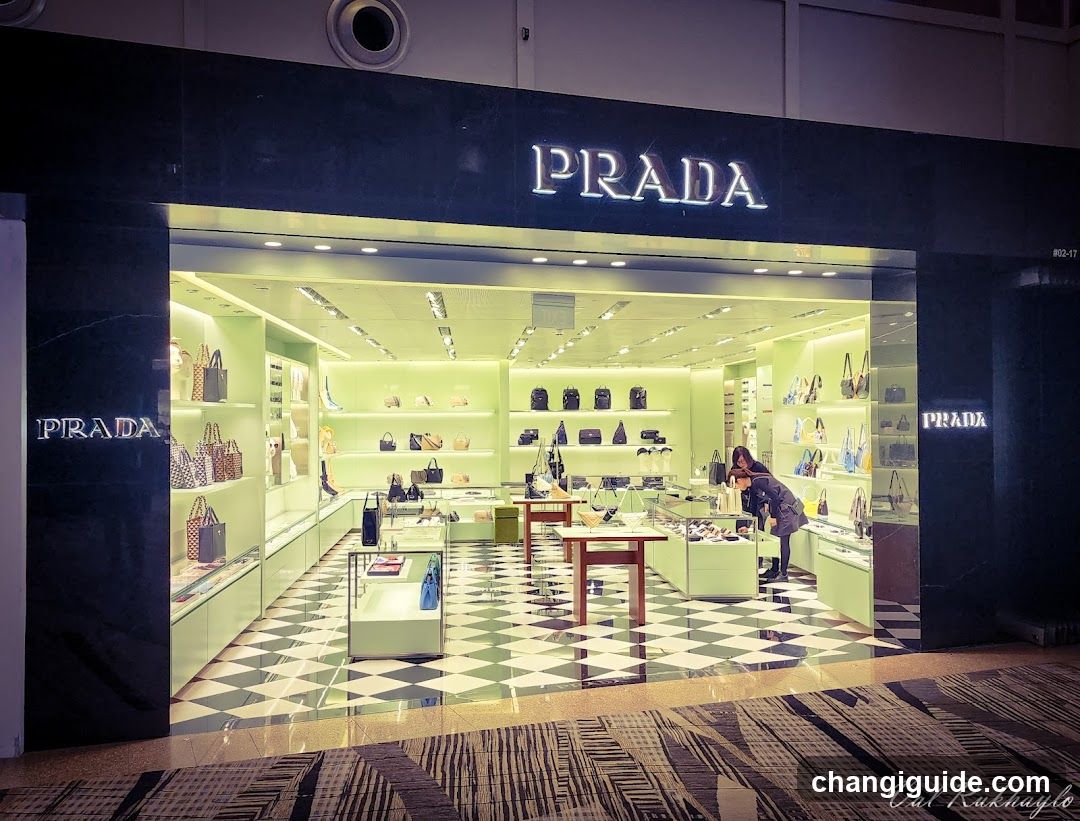 The image shows the exterior of a Prada store with illuminated signage on the facade. The entrance features glass doors and windows displaying various handbags and accessories inside. A person is visible near the entrance, possibly entering or exiting the store.