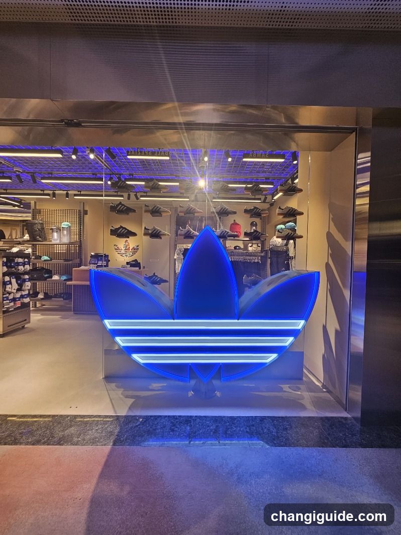 A brightly lit storefront featuring a large, glowing blue Adidas trefoil logo on the glass facade. Inside, shelves display various shoes and apparel under purple accent lighting. The entrance is framed by metallic panels, and the interior reveals organized product displays.