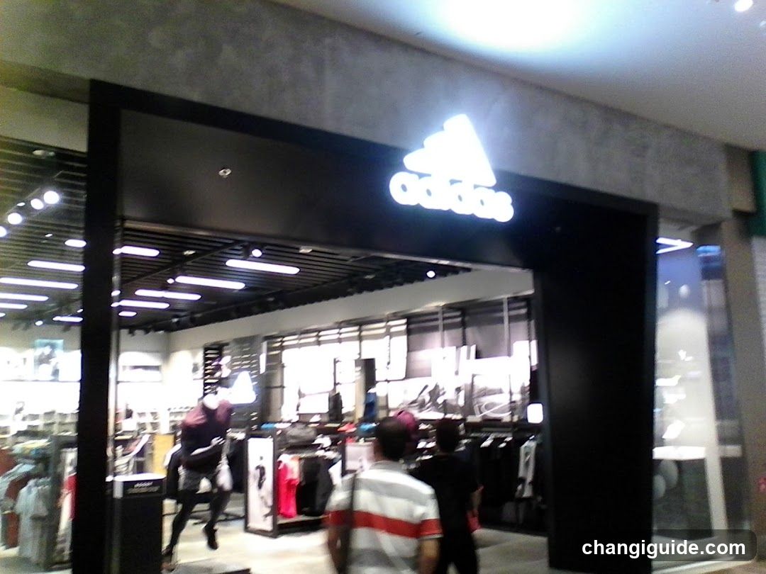 The photo shows the exterior entrance of an Adidas store, featuring a prominent illuminated logo above the doorway. The storefront is modern with large glass windows and doors, allowing visibility into the brightly lit interior where clothing racks and merchandise are displayed. Several people can be seen browsing inside.