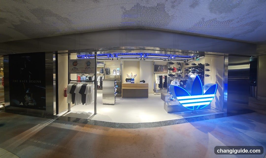 A modern retail storefront featuring a large illuminated Adidas trefoil logo on the right side of the entrance. The store interior is visible through glass doors, displaying clothing racks with shirts and pants, shelves with shoes, and a wooden counter at the center. A promotional poster on the left wall reads 'THE RACE BEGINS'. Blue LED lighting accents the ceiling and display areas.