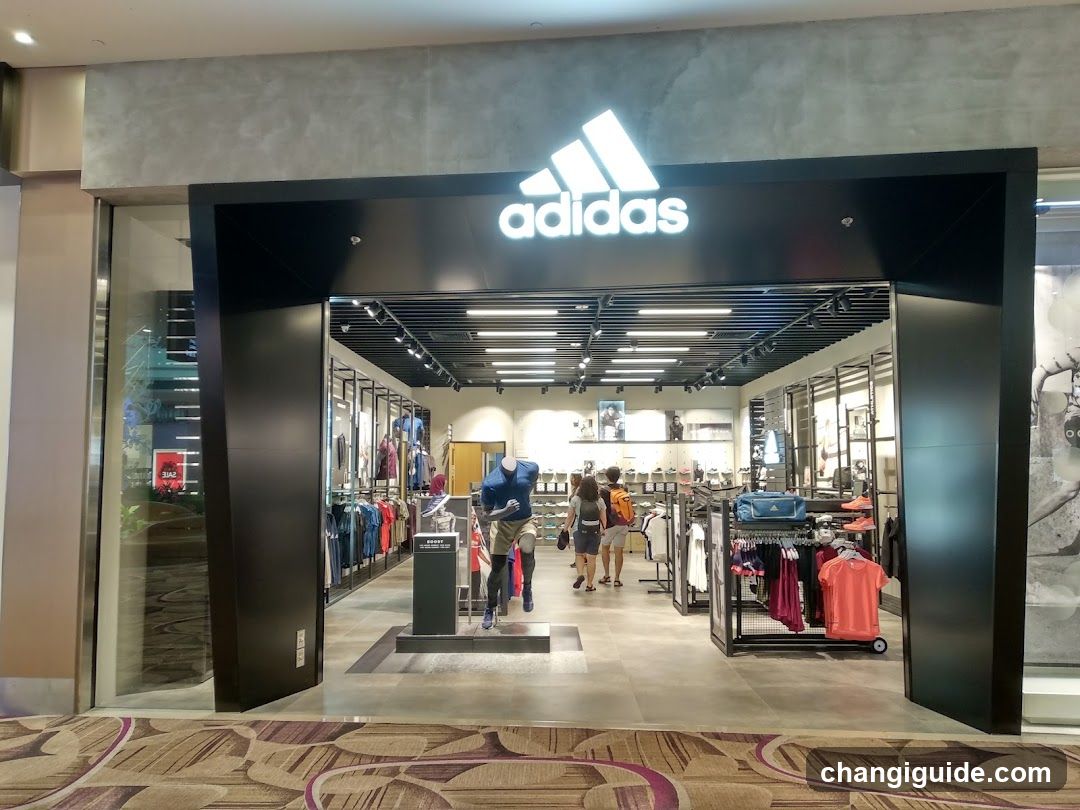 A modern Adidas storefront with a large illuminated logo above the entrance. The store features glass doors, metal shelving units displaying clothing and accessories, and mannequins inside. Two people are walking near the entrance.