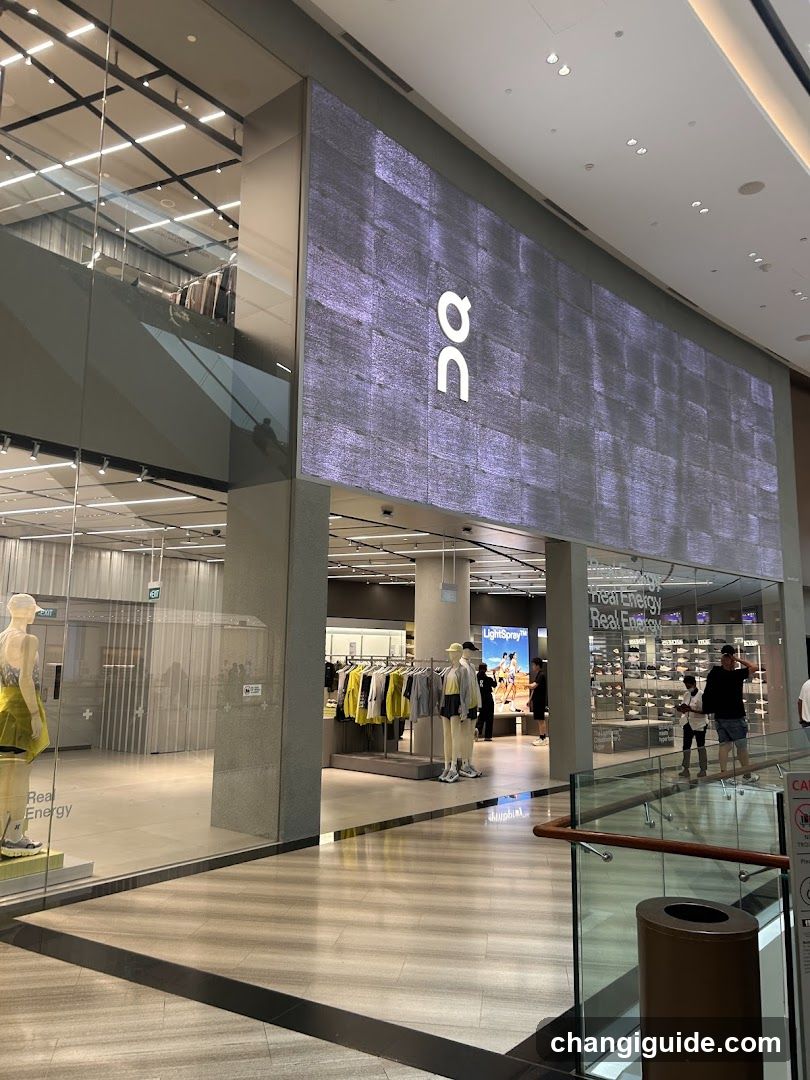 The image shows a modern retail store entrance featuring a large, illuminated sign with the letters 'n' and 'a' vertically aligned on a textured purple background. The storefront is made of glass, allowing visibility into the interior where clothing racks and mannequins are displayed. A few people can be seen inside the store, browsing merchandise. The floor outside has a light-colored tile pattern, and there is a trash bin near the entrance.