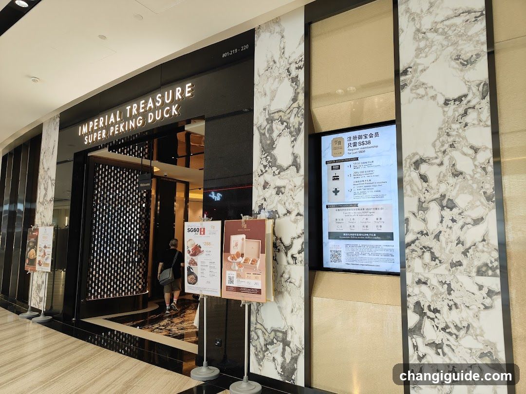 The image shows the exterior entrance of a shop named 'IMPERIAL TREASURE SUPER PEKING DUCK'. The storefront features black and white marble cladding with illuminated signage above the entrance. There are promotional displays in front, including a menu board showing dishes like duck and rice, and another poster advertising membership benefits. A person is seen entering the store.