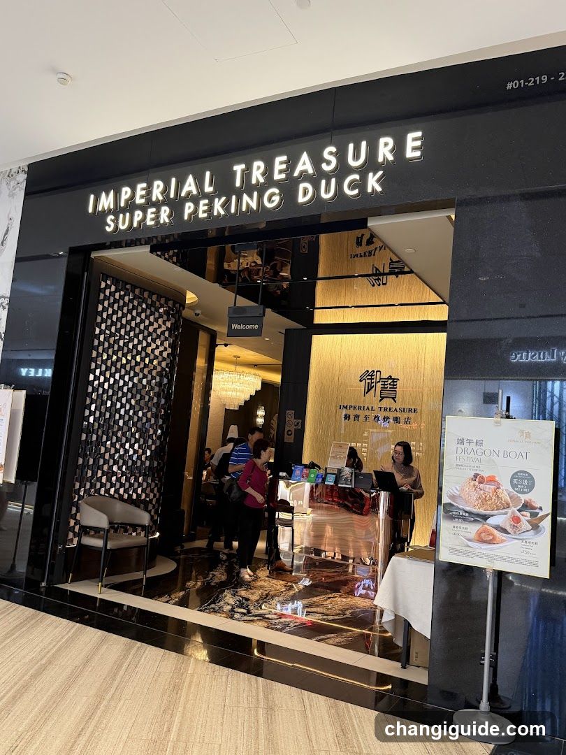 The image shows the storefront of Imperial Treasure Super Peking Duck, a restaurant with illuminated signage above the entrance. The facade features black and gold accents, with a decorative screen on the left side. Inside, customers are visible near the counter area.