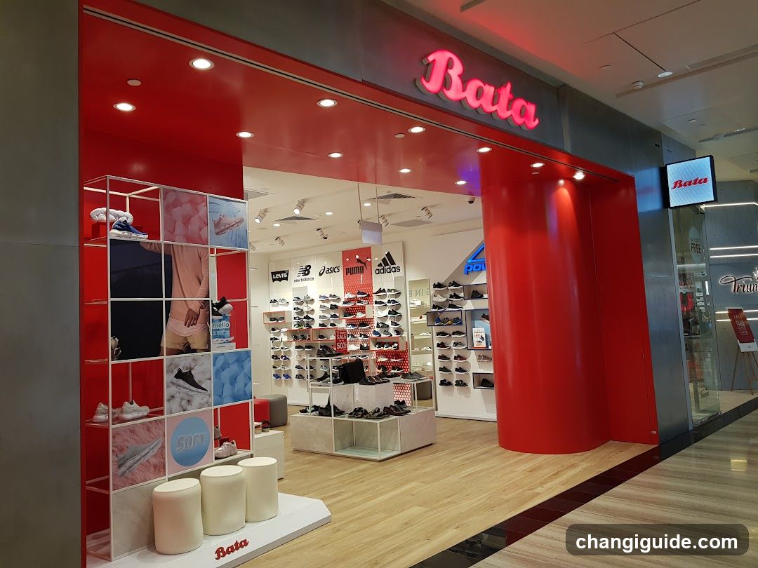 The image shows the storefront of a Bata shoe store, featuring a bright red facade with illuminated 'Bata' signage above the entrance. Inside, shelves display various shoes from brands like New Balance, Asics, Puma, and Adidas. A promotional standee is visible near the entrance.