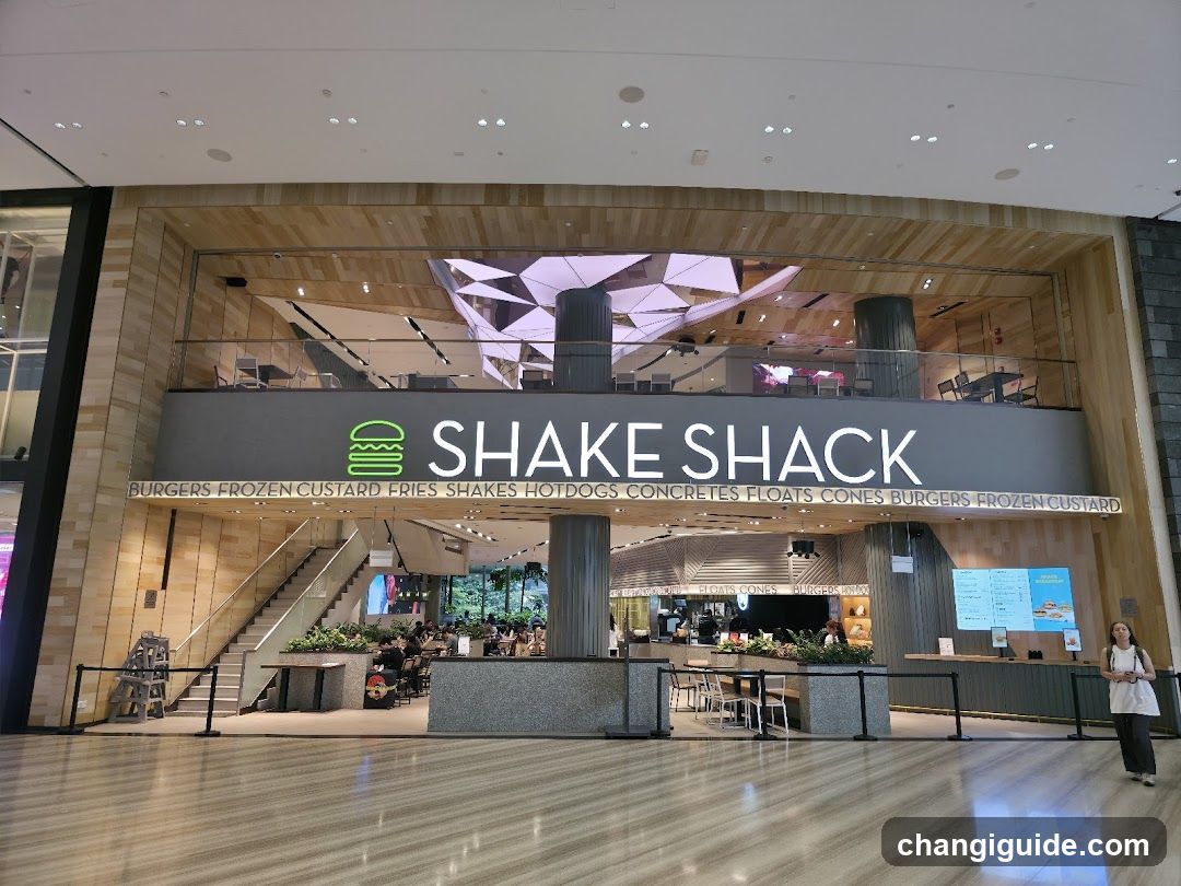 A modern Shake Shack storefront with a large sign displaying the brand name and logo. The entrance features glass doors, wooden paneling, and a visible interior seating area.