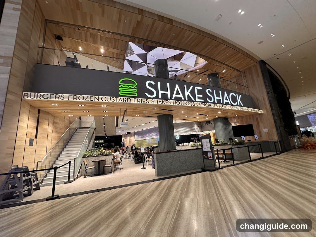 A modern Shake Shack storefront with a large illuminated sign displaying the brand name and logo. The entrance features a staircase leading up to the dining area, surrounded by wooden paneling and glass railings. Inside, customers are seated at tables enjoying food.