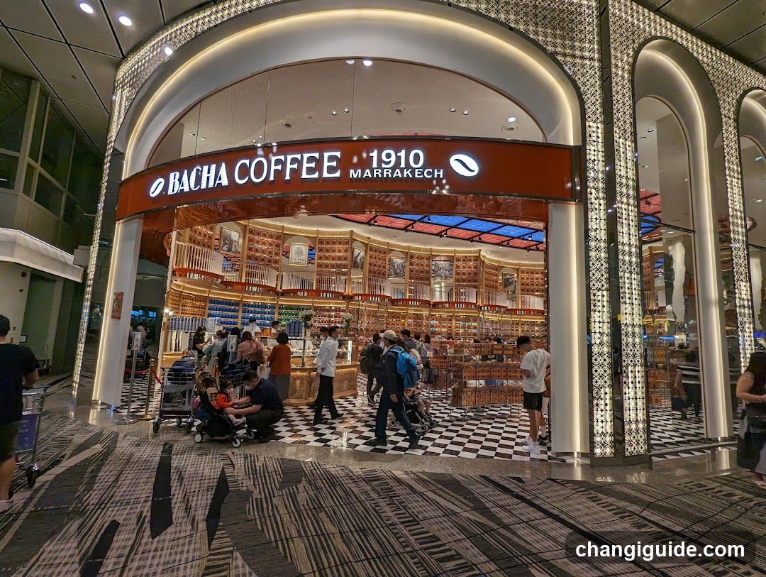 The image shows the exterior entrance of a Bacha Coffee store, specifically the '1910 Marrakech' branch. The storefront features an arched design with decorative columns and a prominent sign displaying the brand name and location. Inside, customers are browsing through shelves filled with coffee products, and the interior has a checkered floor pattern.