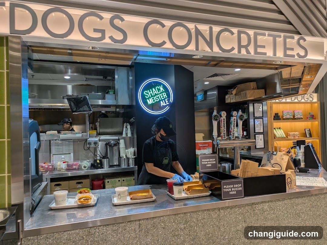A food stall with a sign reading 'DOGS CONCRETES' above the entrance. Inside, staff in black uniforms and masks prepare food at a stainless steel counter. A neon sign displays 'SHACK MEISTER'S ALE'. Trays of burgers are visible on the counter, along with signs instructing customers to drop buzzers here.