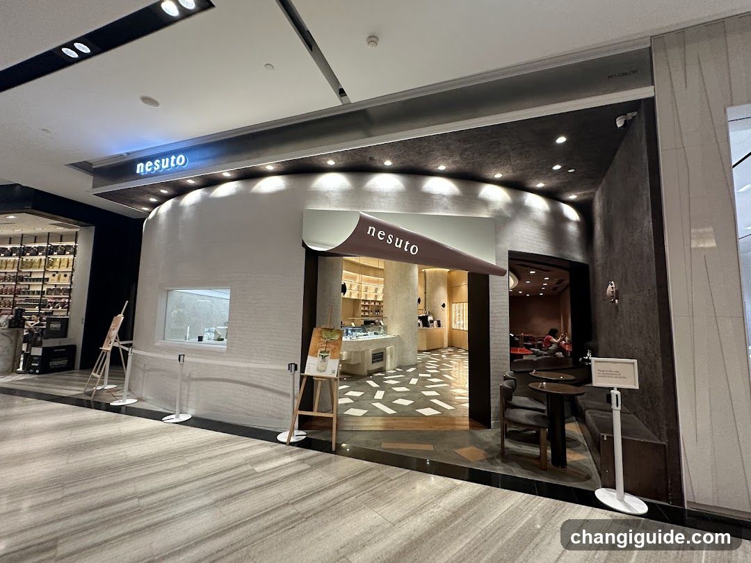A modern shop front with a curved white facade and dark ceiling accents. The nesuto logo is visible on the awning above the entrance and mounted on the building's exterior wall. Inside, there are wooden shelves displaying products, a counter area, and seating with tables and chairs. A small signboard stands near the entrance.