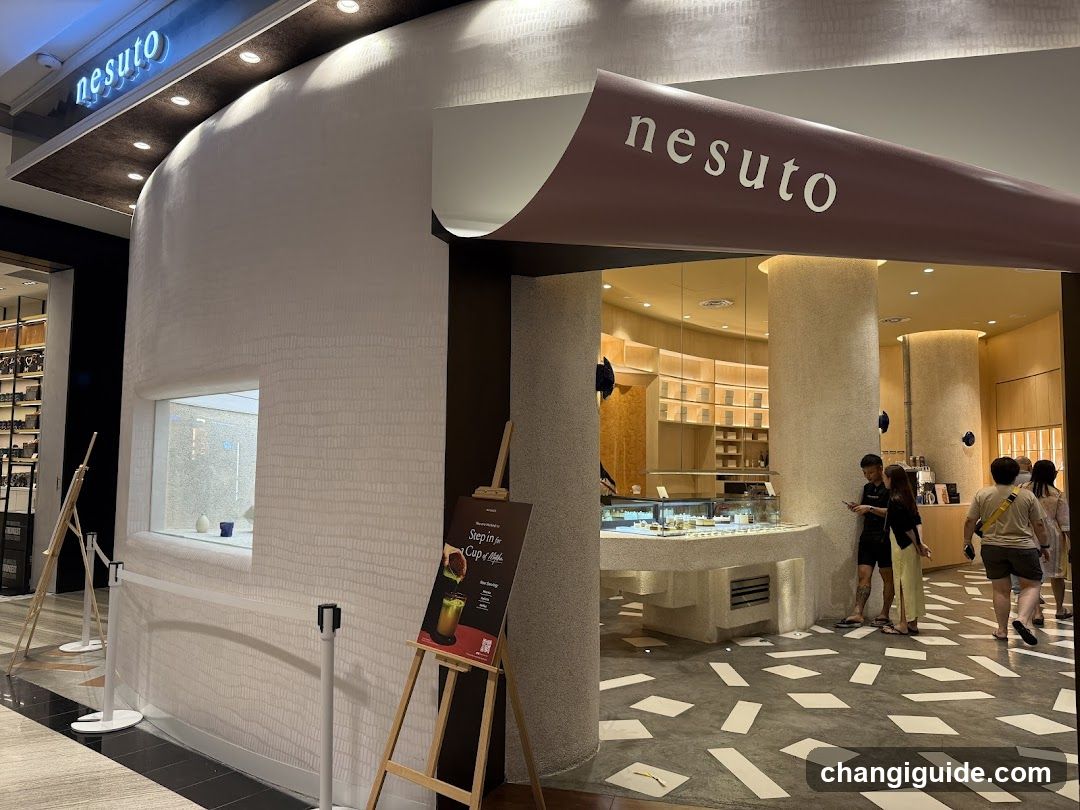 A modern, curved white facade with 'nesuto' signage above the entrance and on a maroon awning. Inside, customers browse near glass display counters under warm lighting.