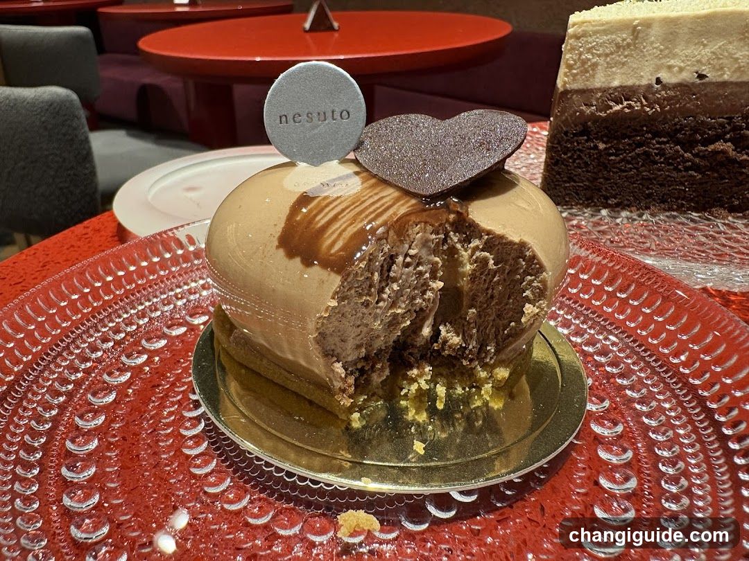 A close-up of a chocolate dessert on a red textured plate, featuring a glossy chocolate glaze and a heart-shaped decoration. A bite has been taken out, revealing the creamy interior. In the background, there is another slice of cake and a red table.