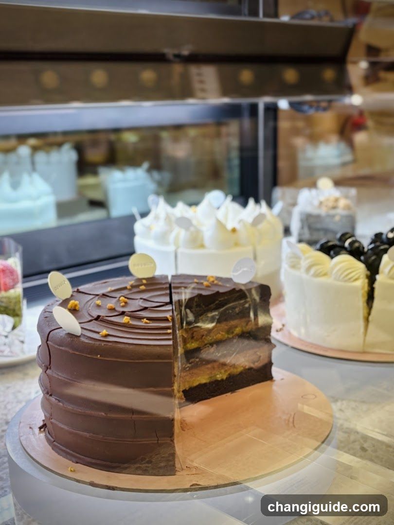 A close-up view of a chocolate cake with a slice removed, revealing layers inside. The cake is decorated with white and gold accents on top. In the background, other cakes are visible in a display case.