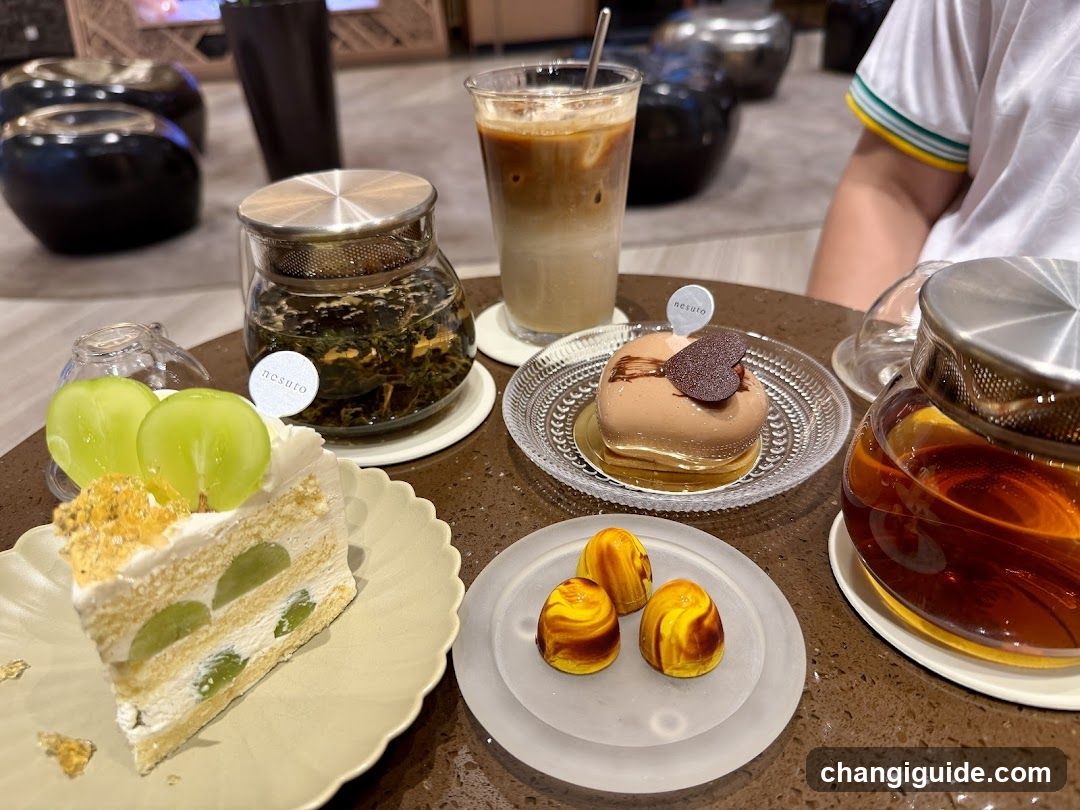 A table setting featuring a slice of layered cake with green grapes, three small pastries on a white plate, a glass jar of loose-leaf tea, an iced coffee drink, and another glass jar containing amber-colored liquid (likely syrup or honey). A person's arm is partially visible in the background.