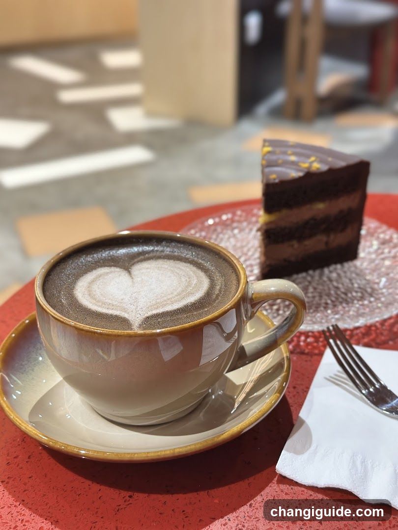 A cup of coffee with heart-shaped latte art sits on a saucer, next to a slice of layered chocolate cake on a red table.