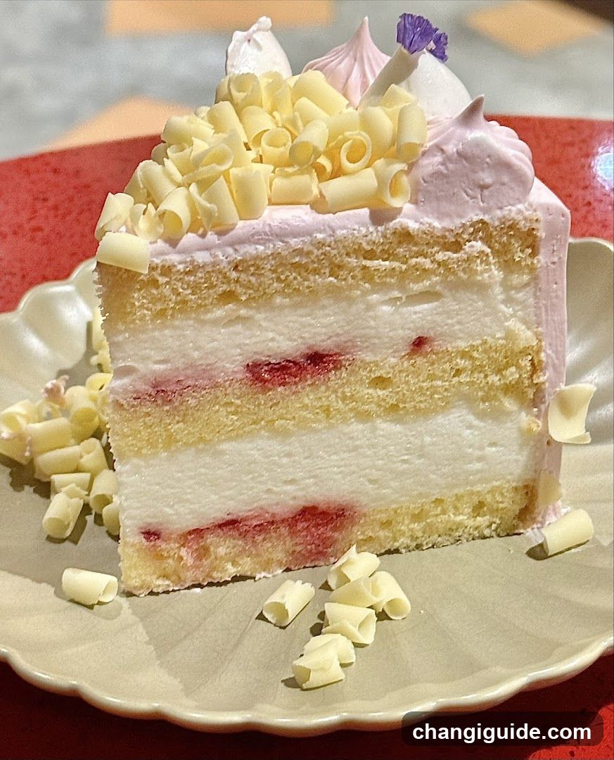 A slice of layered cake with vanilla sponge, cream filling, and strawberry jam layers. Topped with white chocolate curls, pink frosting rosettes, and a purple flower garnish. Served on a beige scalloped plate with scattered white chocolate shavings.