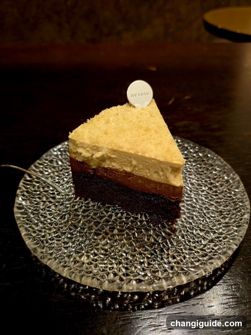 A triangular slice of multi-layered dessert sits centered on a clear, etched glass plate with a scalloped rim. The cake features alternating layers of dark chocolate sponge and lighter brown mousse or cream filling, topped with a thick layer of pale yellow whipped cream or custard. A small white circular topper with the word 'nesu' is placed at the peak of the cake. A thin metal fork rests partially under the left side of the plate. The background is dimly lit, showing a dark wooden surface and blurred interior elements.
