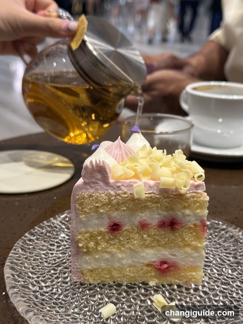 A slice of layered cake with pink frosting, white chocolate shavings, and raspberry filling sits on a textured glass plate. A hand pours tea from a glass teapot into a small cup nearby. Another coffee cup is visible in the background.