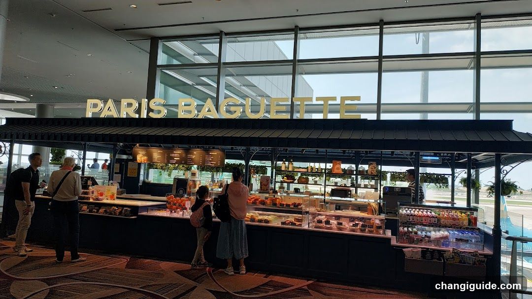 A Paris Baguette storefront with large glass windows and a prominent sign above the counter. Several customers are browsing inside, and various baked goods and beverages are displayed on shelves.