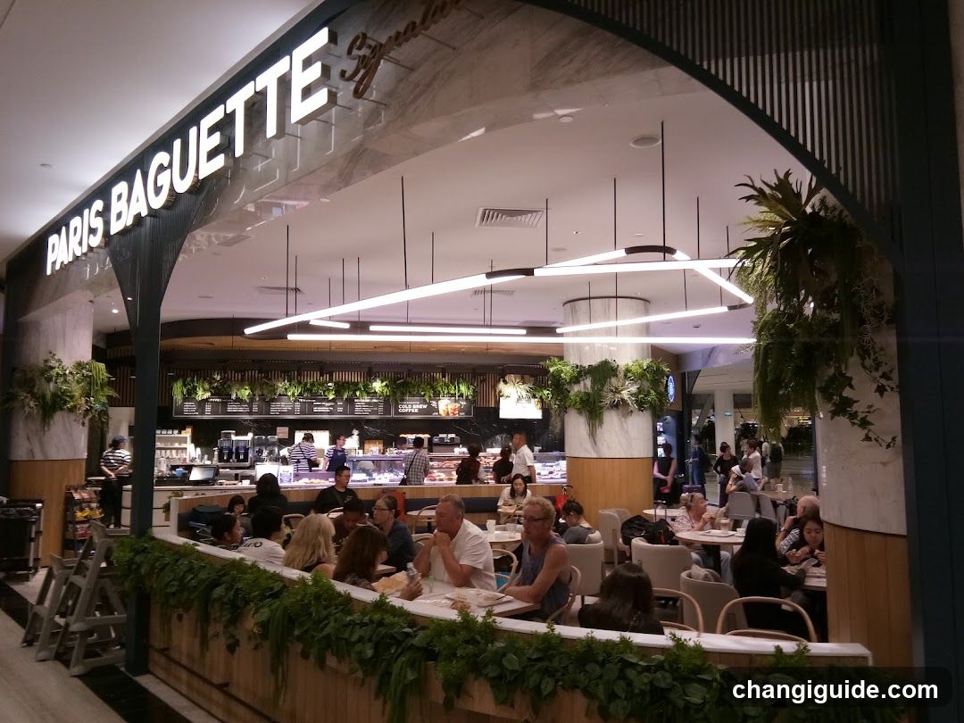 The image shows the storefront of a Paris Baguette bakery, with its name prominently displayed in white letters on a dark background. The entrance is framed by an arched design, and there are green plants adorning the area. Inside, customers can be seen seated at tables, enjoying their meals. The interior features modern lighting fixtures and a counter where staff members are working.