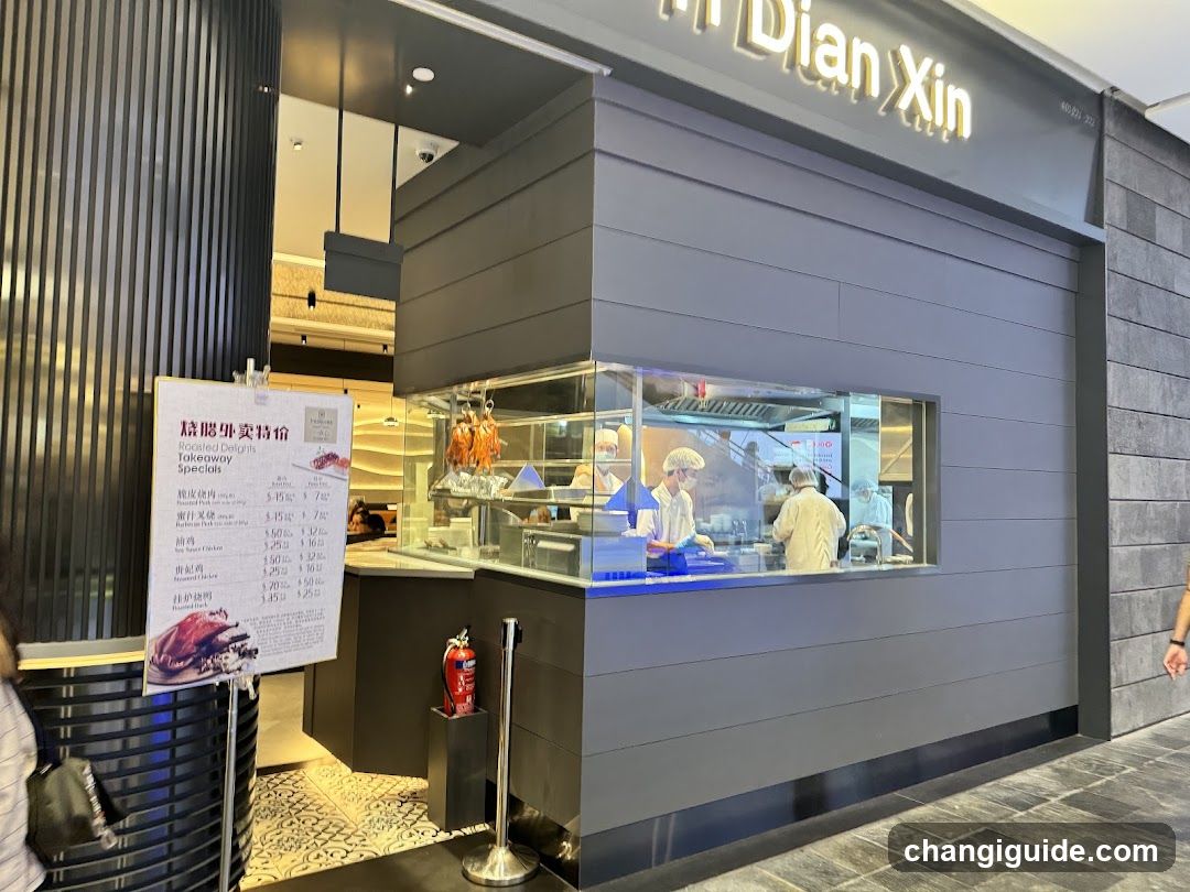 A modern shop front with a gray facade and illuminated signage reading 'Dian Xin'. Inside, staff in white uniforms are preparing food behind a glass counter. A menu board is visible on the left side of the entrance.