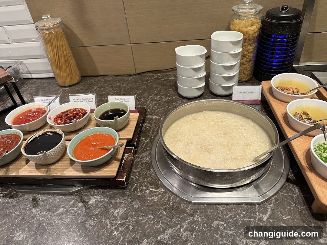 A breakfast buffet setup featuring a large pot of congee (rice porridge) in the center, surrounded by various condiments and toppings including sauces, chopped vegetables, and garnishes. Stacks of white bowls are neatly arranged nearby, along with jars of pasta and other ingredients.