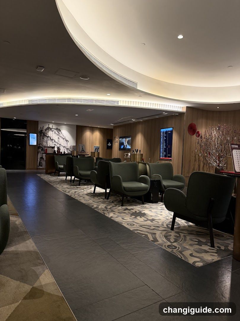 A modern, well-lit lobby or waiting area featuring dark green armchairs arranged on a patterned rug over polished tile flooring. A curved ceiling design with recessed lighting dominates the space. In the background, there is a wall mural depicting an architectural scene, and digital screens displaying flight information or schedules are mounted on wooden-paneled walls. Decorative elements include red circular ornaments and a stylized tree arrangement near the right side. No visible menus or shop fronts are present.