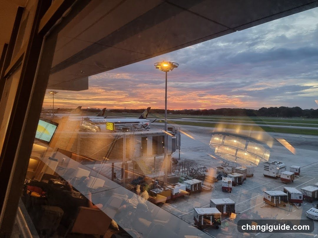 A view from inside an airport terminal looking out onto the tarmac during sunset. The sky is filled with vibrant orange and pink hues near the horizon, transitioning to darker blues above. Several airplanes are parked at gates, connected by jet bridges. Ground service vehicles and equipment are visible on the apron. Reflections of interior lights and possibly passengers or staff can be seen on the glass window in the foreground.