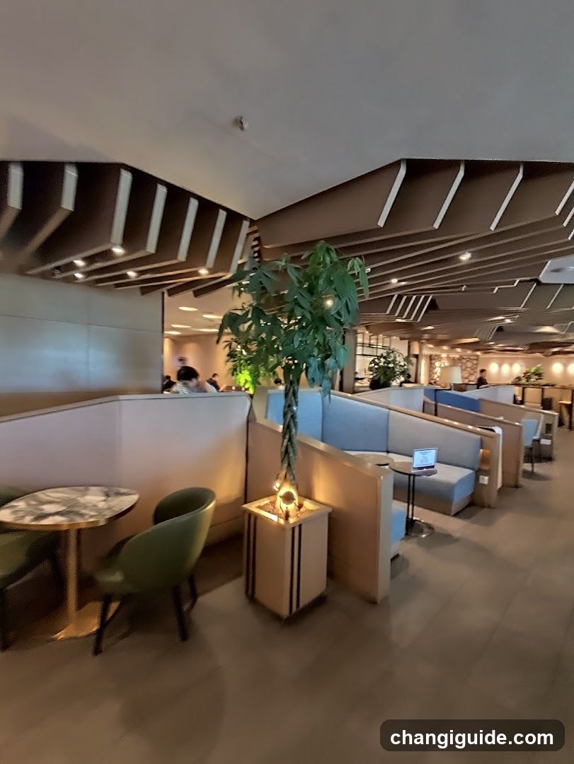 Interior view of a modern lounge or café with wooden ceiling beams, booth seating in light blue and beige tones, green accent chairs, marble-topped tables, potted plants, and ambient lighting. A laptop is visible on one table. No signage, menus, or exterior storefront elements are present.