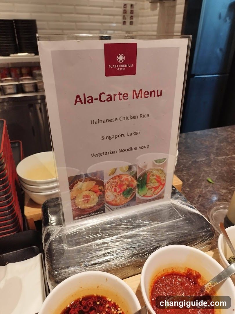 A close-up view of a food service area featuring an 'Ala-Carte Menu' sign listing Hainanese Chicken Rice, Singapore Laksa, and Vegetarian Noodles Soup with corresponding images. In the foreground are bowls containing red chili sauce and other condiments.