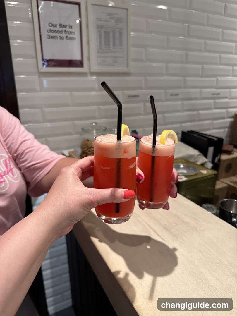 A person in a pink shirt holds two glasses of orange-colored beverages with straws and lemon slices, clinking them together. The background shows a white tiled wall with framed notices including one stating 'Our Bar is closed from 3am to 8am'. A wooden countertop and kitchen equipment are visible behind the drinks.