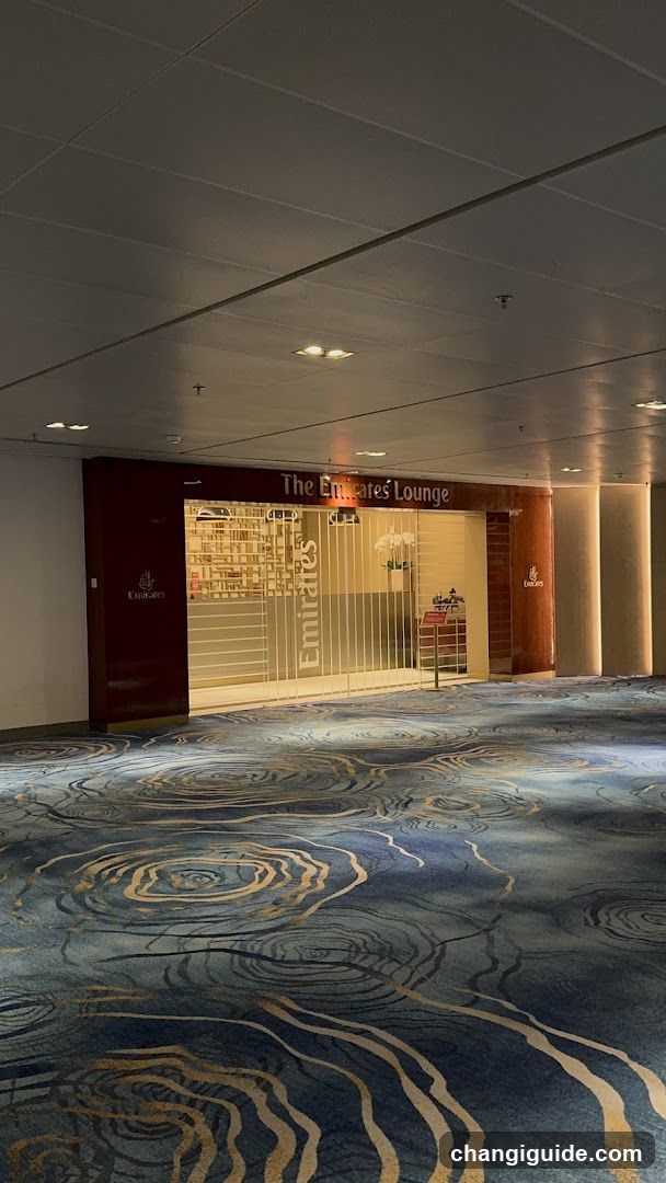The image shows the interior of The Emirates Lounge, featuring a patterned blue carpet with gold swirls leading to a glass entrance. Inside, there is a reception desk and shelves displaying various items. The lighting is warm, creating an inviting atmosphere.
