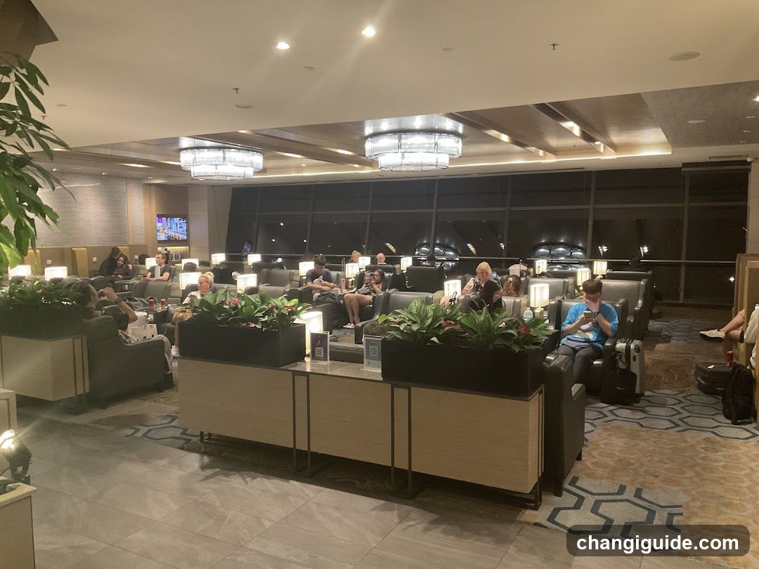 A spacious airport lounge with rows of comfortable armchairs, each equipped with personal lighting and small tables. Passengers are seated, some reading or using devices. Large windows overlook a dark exterior at night. Decorative plants in planters line the center aisle. A television screen is visible on the left wall.