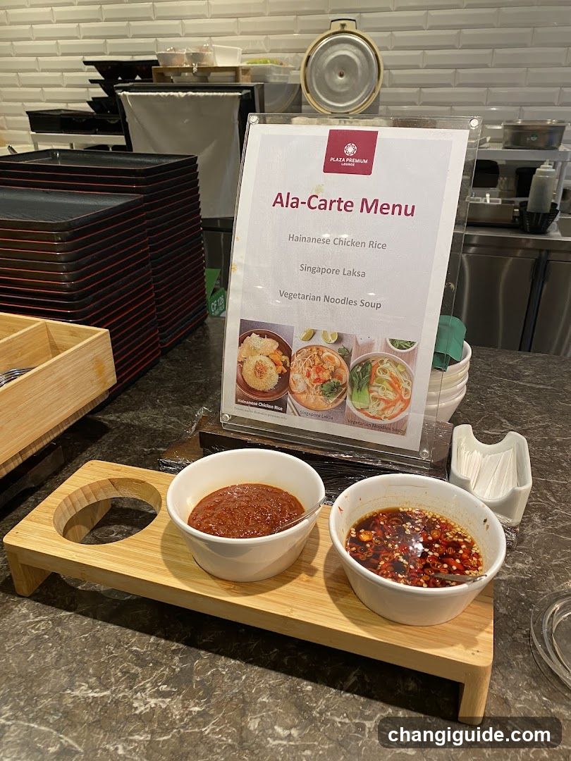 A close-up view of a dining setup featuring two small white bowls filled with sauces—one red chili oil and one dark brown sauce—placed on a wooden tray. Behind them is an 'Ala-Carte Menu' sign listing Hainanese Chicken Rice, Singapore Laksa, and Vegetarian Noodles Soup.