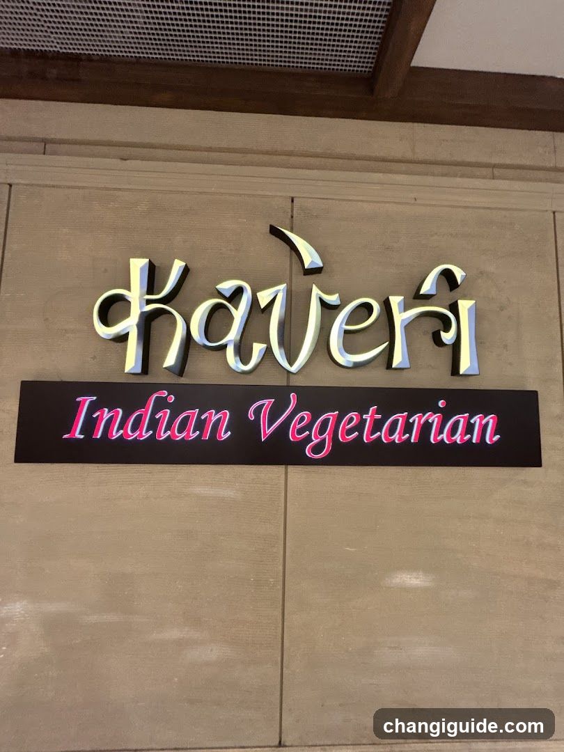 A close-up photo of a shop sign mounted on the exterior wall. The sign features stylized metallic lettering spelling 'kaveri' in Devanagari script above a black rectangular panel with pink neon-style text reading 'Indian Vegetarian'. The background is a light beige tiled wall under an exposed wooden beam and ventilation grille.