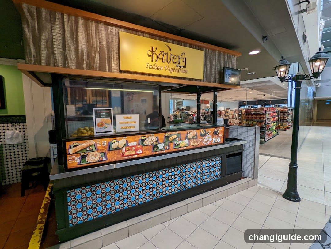 A food stall in a mall with a yellow sign reading 'Kaveri Indian Vegetarian'. The counter features a menu board displaying various dishes like bread, samosas, and curries. Decorative blue tiles line the base of the stall.