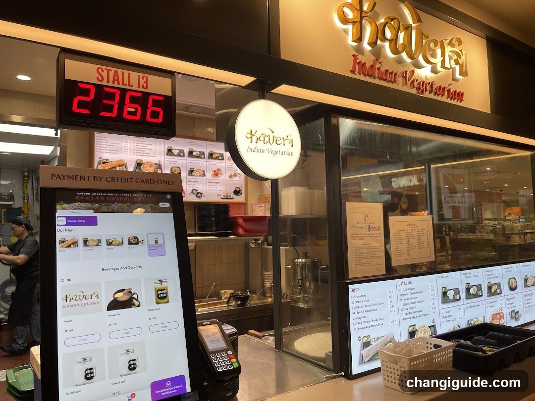 A food stall in a mall with illuminated signage reading 'Kaveria Indian Vegetarian'. A digital display shows 'STALL I3 2366'. A self-service kiosk displays menu options like Yogurt and Smoothies. Inside, staff are visible behind the counter. Menu boards on the wall show various dishes. Payment terminal is present at the front.