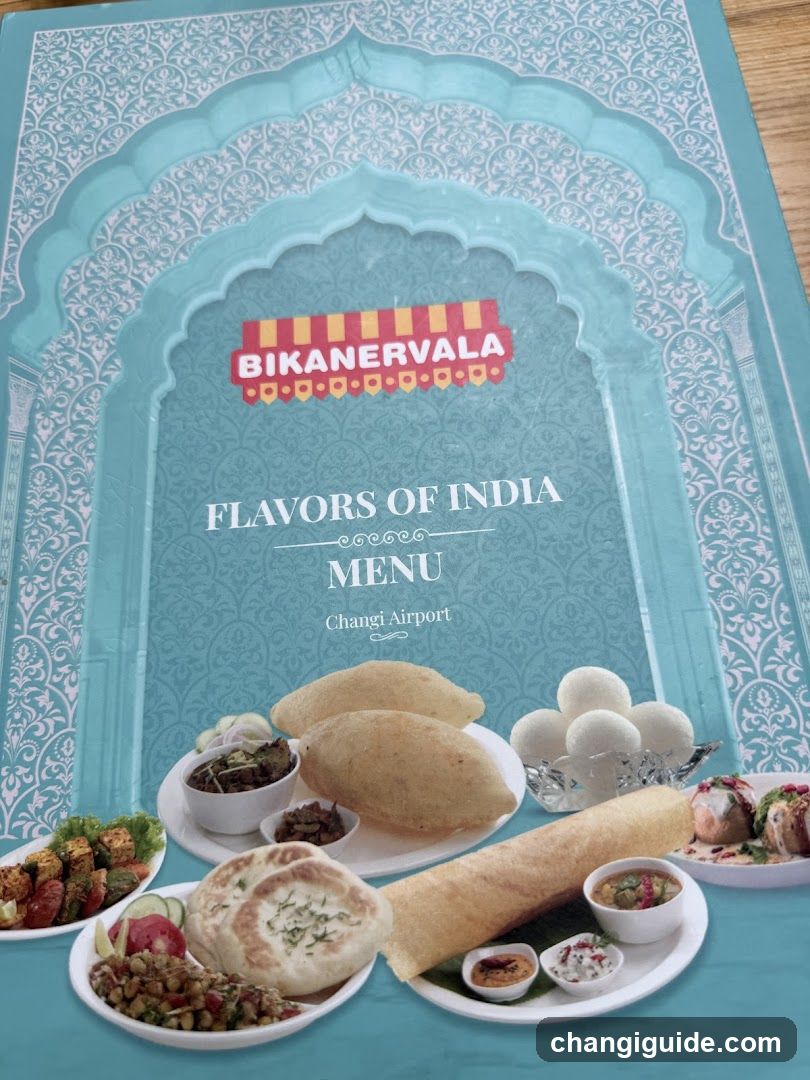 A printed menu board featuring an ornate blue arch design with the BikanerVala logo and title 'Flavors of India Menu Changi Airport'. The bottom half displays a variety of Indian dishes including samosas, parathas, curries, and sweets arranged on white plates.
