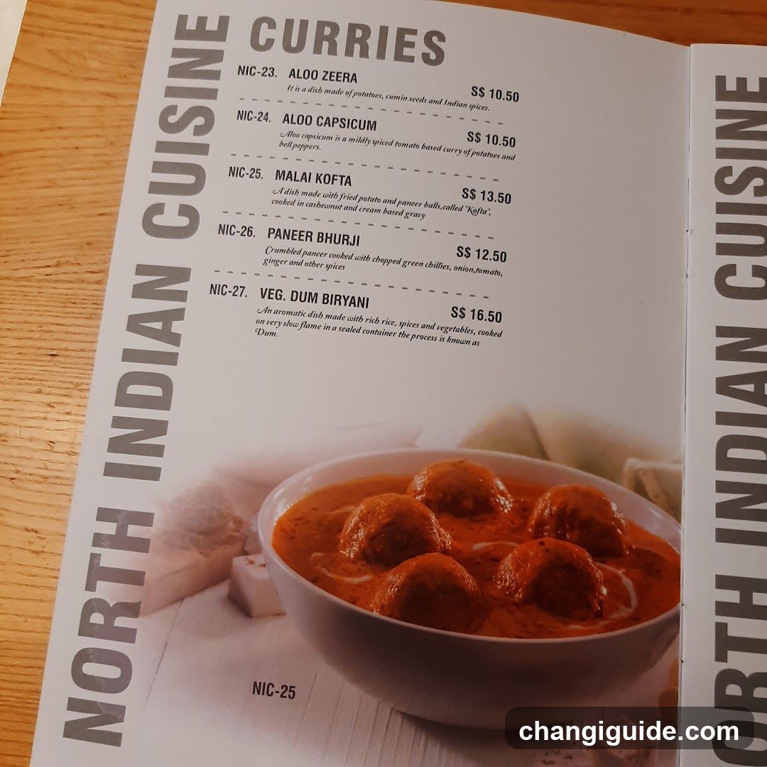 A printed menu page featuring a list of North Indian curry dishes with descriptions and prices, alongside a photo of a bowl of meatballs in red sauce.