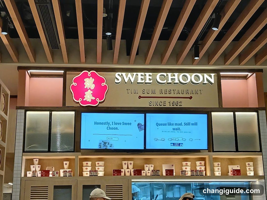 The image shows the storefront of Swee Choon Tim Sum Restaurant, featuring a prominent sign with 'SWEE CHOON TIM SUM RESTAURANT SINCE 1962' and a red emblem with Chinese characters. Below the sign are two digital screens displaying humorous messages about queueing. The lower section displays neatly arranged takeaway containers on shelves.