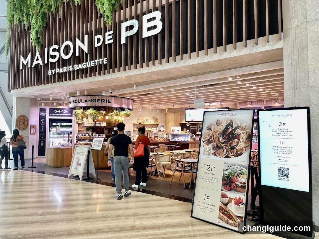 The image shows the storefront of Maison de PB, a bakery and café chain. The sign is prominently displayed on the building facade with vertical wooden slats. Inside, customers are browsing pastries and seated at tables. There are menu boards outside displaying food items like pasta and sandwiches.