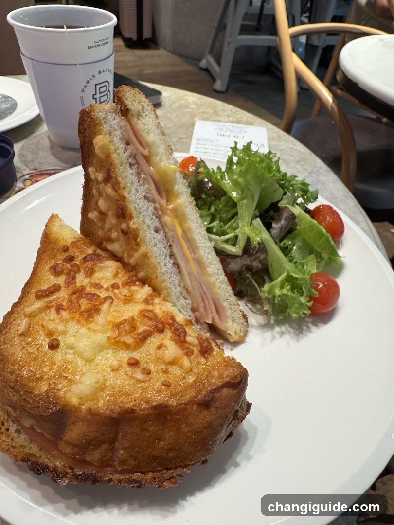A plate featuring a golden-brown grilled sandwich cut diagonally, revealing layers of ham and cheese inside. Accompanied by a small side salad with mixed greens and cherry tomatoes. A white cup with 'PARIS BAGUETTE' branding sits in the background on a wooden table.